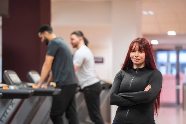 Smiling woman with crossed arms standing confidently in a gym, treadmill background.