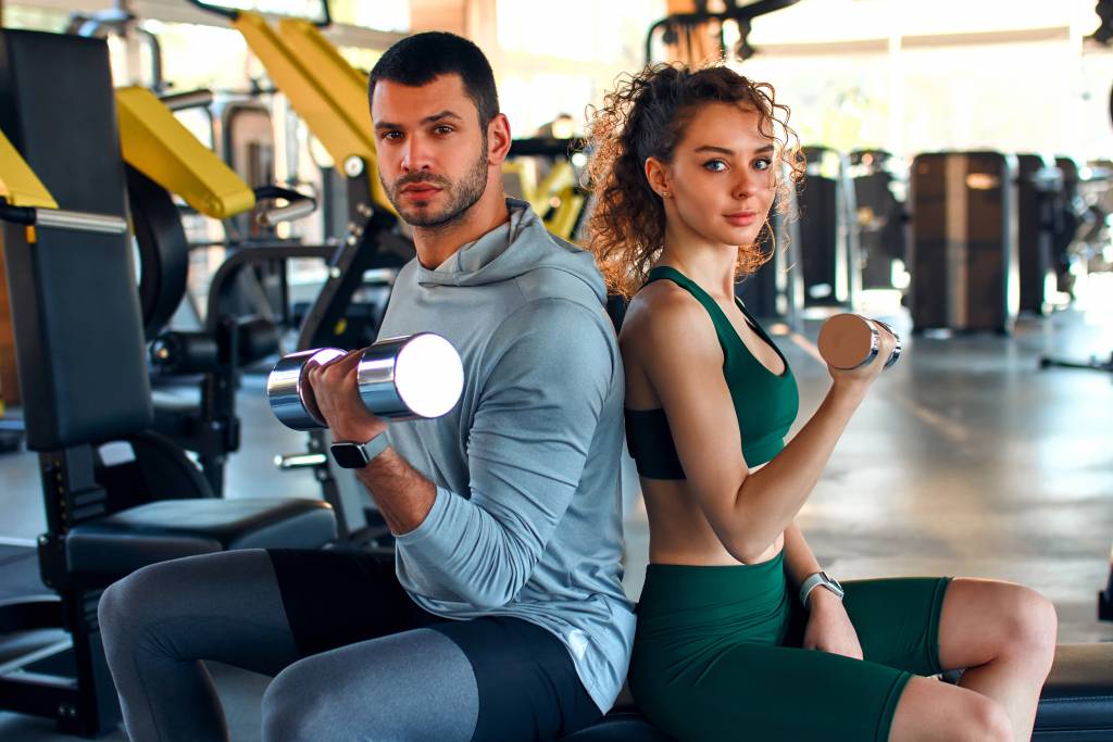 A slender woman in sportswear does exercises with a trainer in the gym with dumbbells on the background of sports equipment. Active healthy lifestyle, sport.